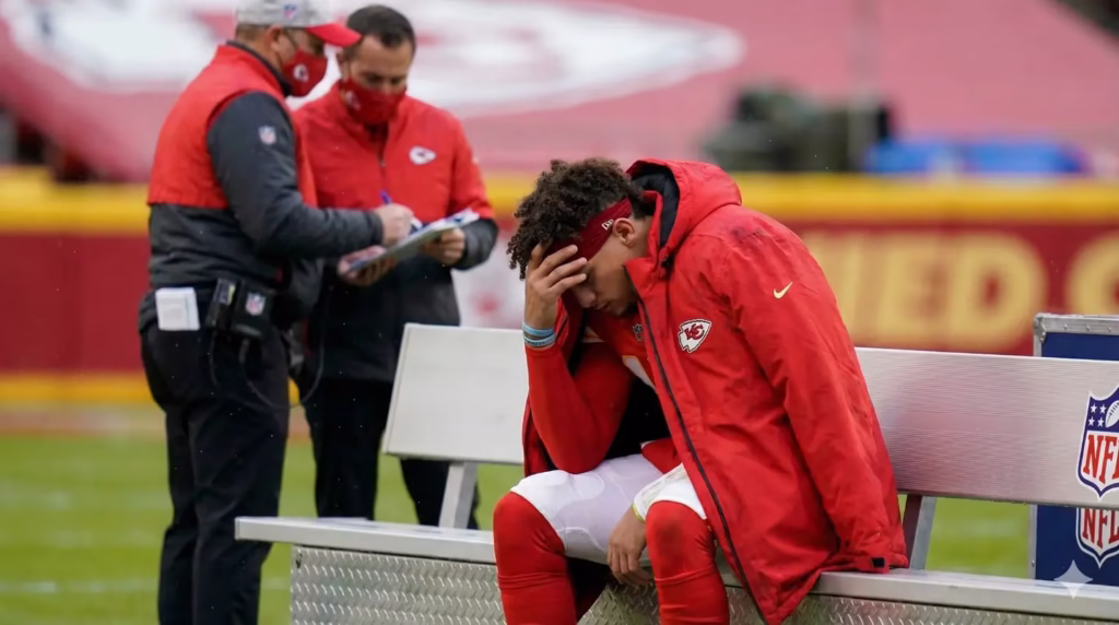 Patrick Mahomes sitting on the sidelines looking disappointed with medical staff in the background.